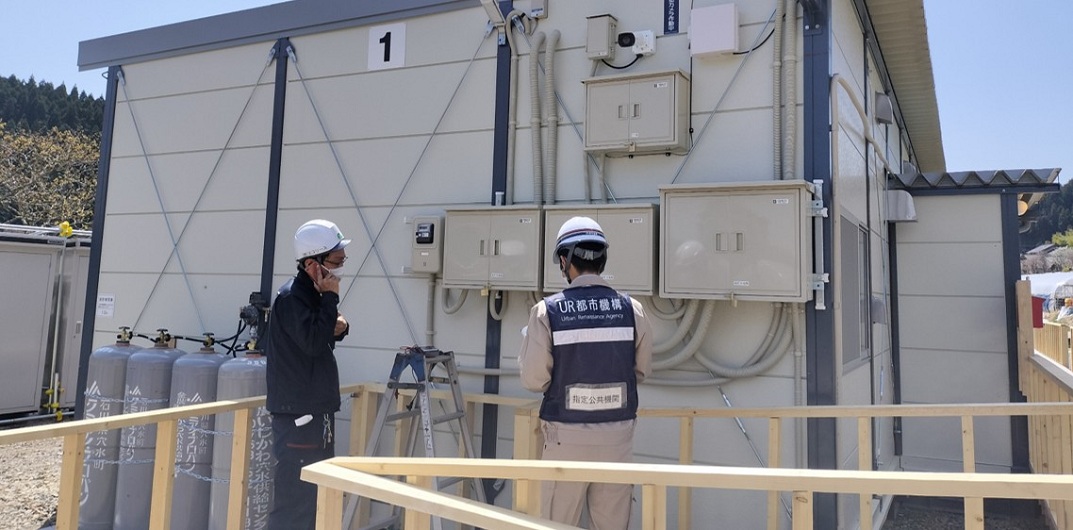 「UR都市機構」と記載されたベストを着用した作業員が、仮設住宅の外壁に設置された電気設備や配管の検査をしている様子。