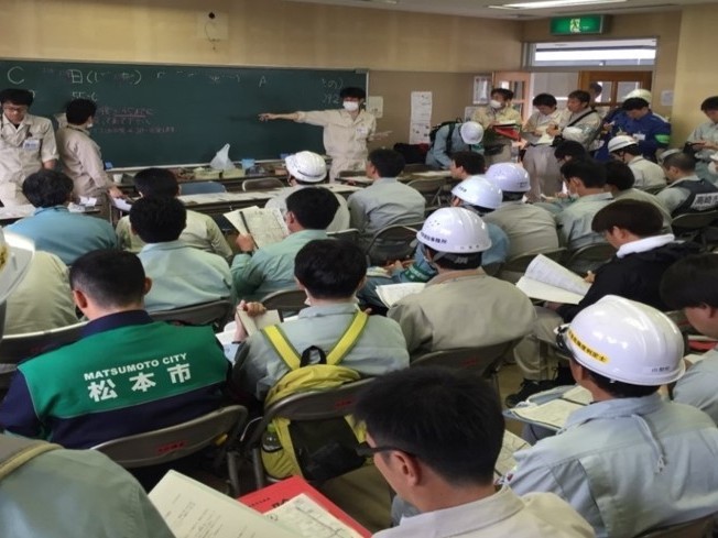 多数の作業員がヘルメットを着用し、黒板に書かれた情報をもとに説明を受けている様子。
