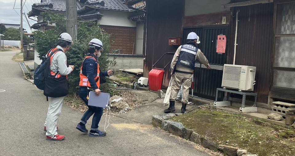 ヘルメットと安全ベストを着用した複数の作業員が、住宅の玄関付近で調査や確認を行っている写真