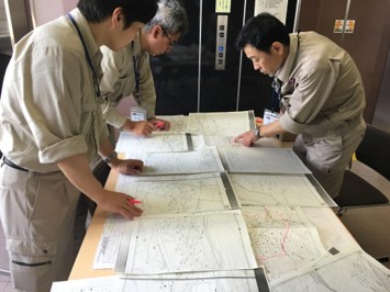 大きな地図や図面が机いっぱいに広げられ、複数の担当者が指示や確認をしている写真