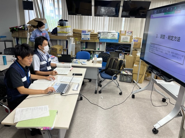 会議室で大型モニターに「3. 調査・判定方法」と表示され、机には資料とノートパソコンが並んでいる。複数の担当者が画面を見ながら業務手順を説明している様子。