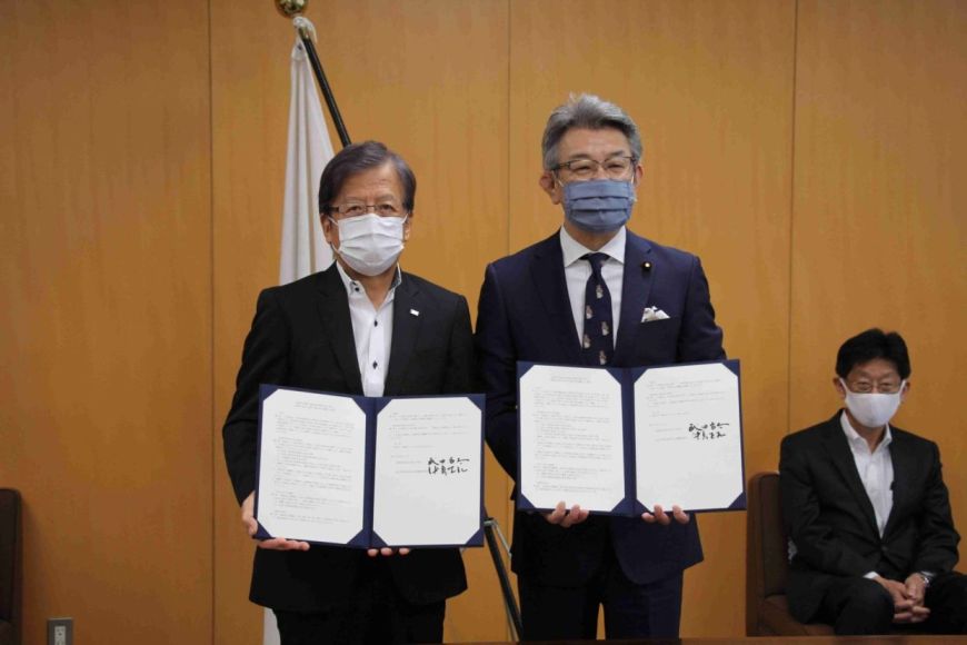 木目調の壁の前で2人が協定書を持って撮影されている場面。書類には署名があり、災害時の支援や連携に関する協定締結の記念写真