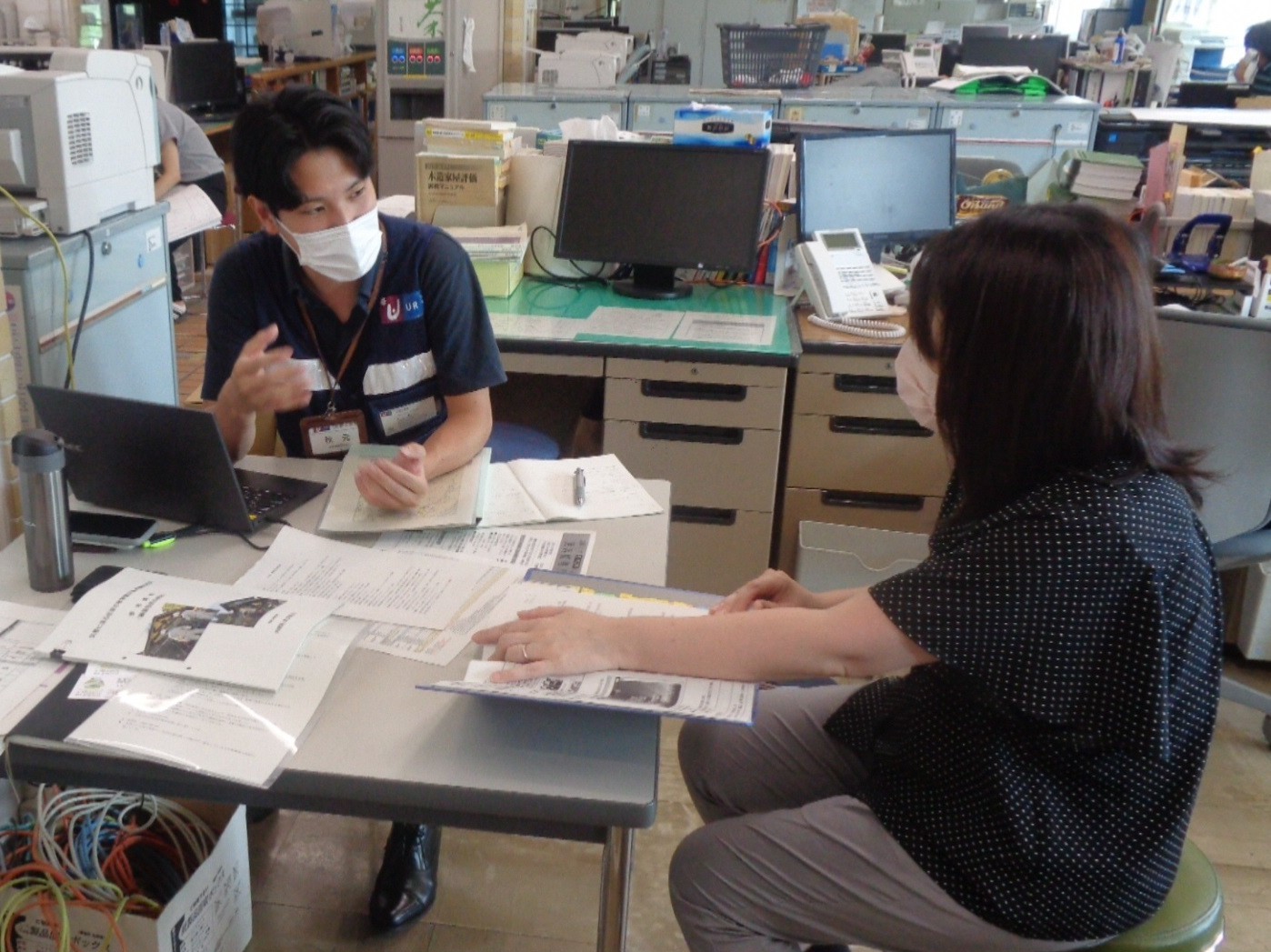 オフィス内で机に資料やノートパソコンが広げられ、2人が対面で業務の説明や確認をしている様子。