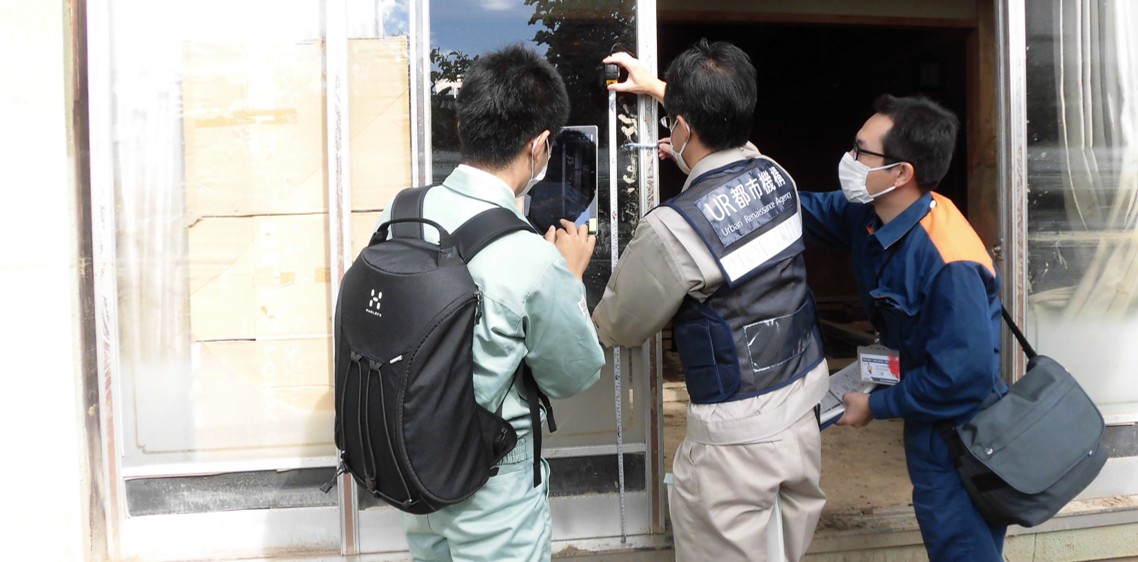 UR都市機構と書かれたベストを着用した調査員が建物の窓枠の損傷状況を確認し、記録している様子。