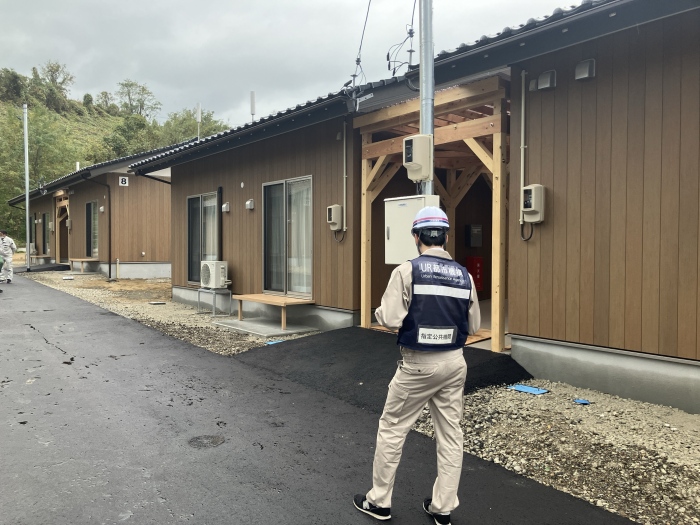 UR都市機構と書かれた服を着た調査員が木造仮設住宅の外観と設備を確認している場面。
