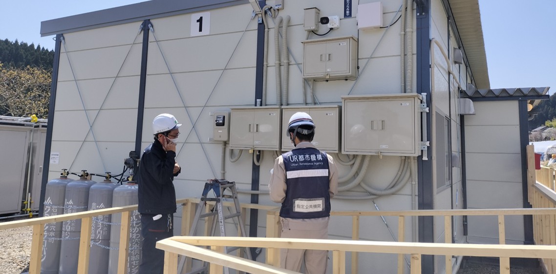 UR都市機構と書かれた服を着た調査員が仮設住宅の外壁に設置された配電盤やガス設備などを点検している様子。