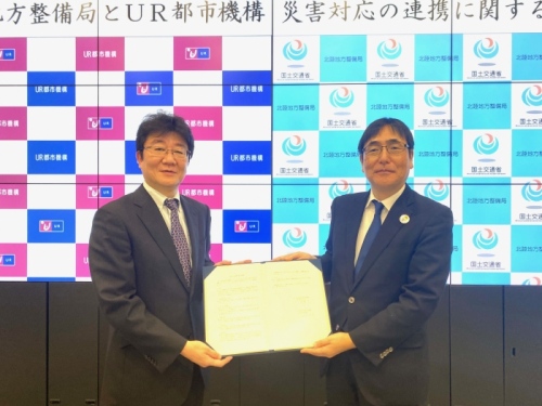 国土交通省北陸地方整備局とUR都市機構のロゴが並ぶ背景の前で、2人が協定書を持って撮影している様子。