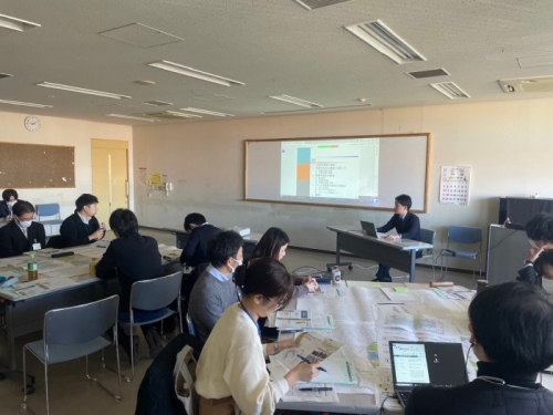 会議室で複数の参加者が資料を広げて議論し、前方では講師がパソコンを使ってスクリーンに資料を投影している様子。
