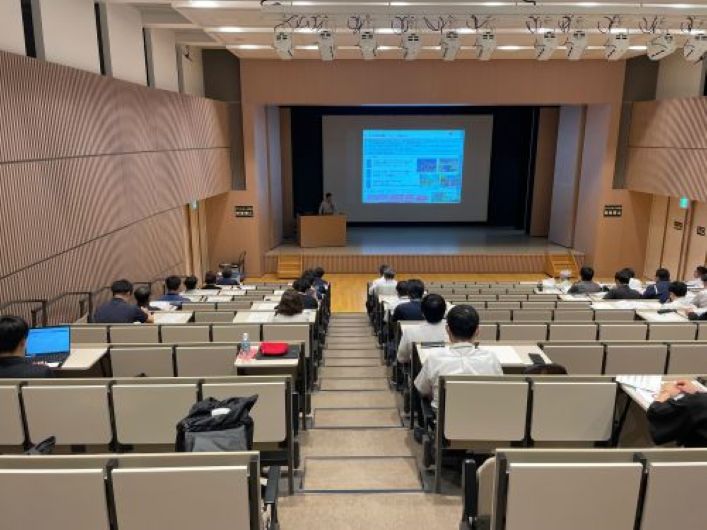 講堂で参加者が前方のスクリーンに映し出されたプレゼンテーションを見ながら講義を受けている様子。