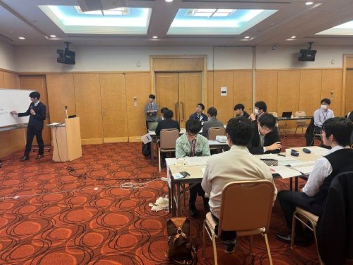 ホテルの会議室で講師がホワイトボードを使って説明し、参加者がテーブルで資料を見ながら話し合っている様子。