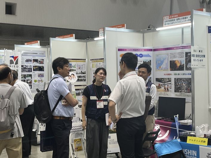 防災関連の展示会場で、複数のパネルや資料が並ぶブースにおいて、来場者とスタッフが立ち話をしながら説明を受けている様子。