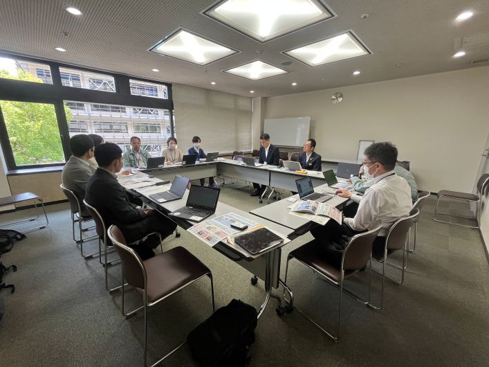 窓のある会議室で複数の参加者が資料やノートパソコンを使って打ち合わせをしている様子。